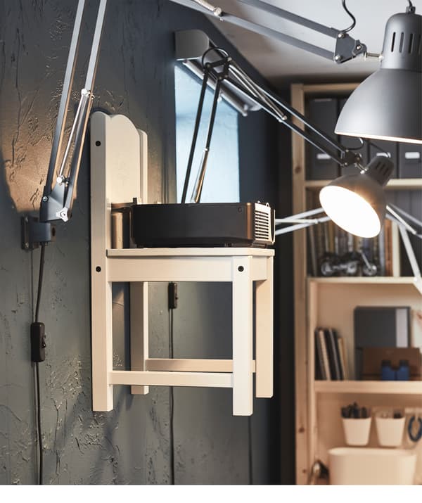Une chaise d’enfant accrochée à un mur par des crochets. La chaise, entourée de lampes de bureaux fixées au mur, supporte un projecteur.