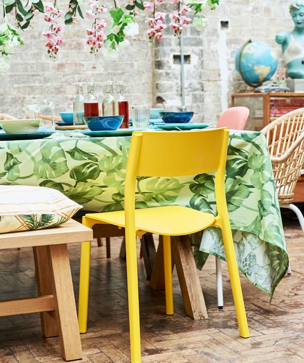 Une chaise en plastique jaune poussée contre une table recouverte d’une nappe à imprimé de feuilles vertes et de vaisselle bleue.