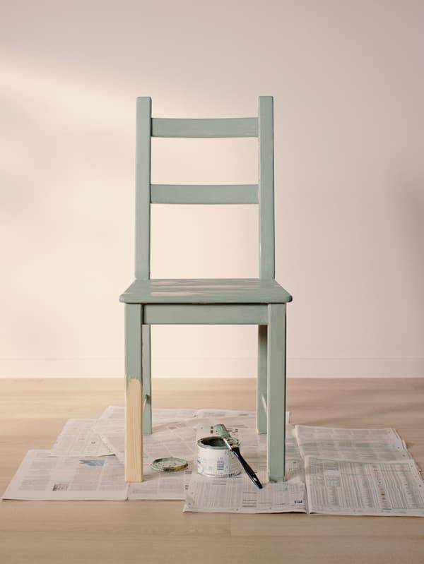 Une chaise IVAR peinte en vert clair posée sur de vieux journaux dans une pièce vide au parquet en bois clair et aux murs rose clair.