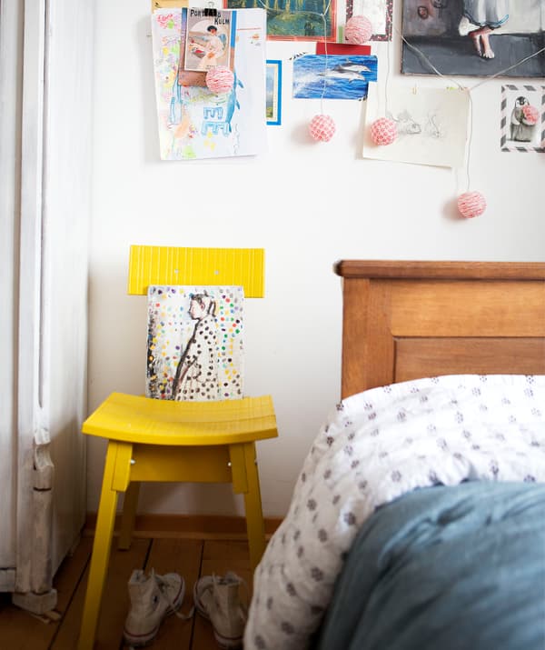 Une chaise jaune contre un mur blanc décoré avec des dessins et des notes à côté d’un lit en bois.