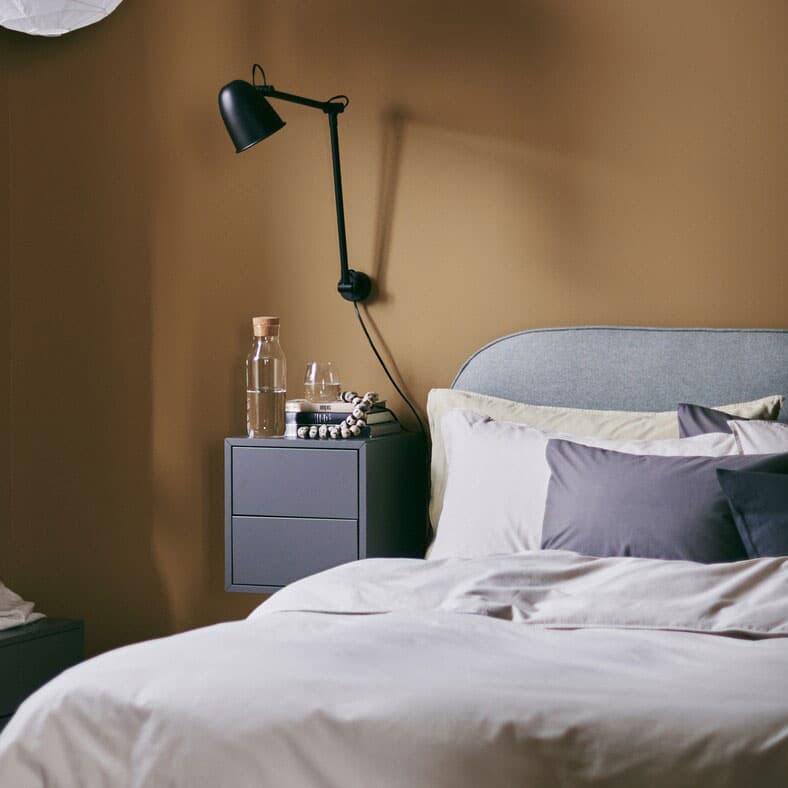 Une chambre à coucher accueillante avec des murs aux tons caramel riches, un lit matelassé VADHEIM, une applique et une table de chevet.