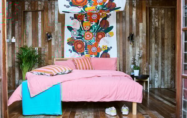 Une chambre à coucher aux murs en lattes de bois avec un lit garni de draps roses, un ciel de lit dans un tissu coloré, et des plantes à côté du lit.