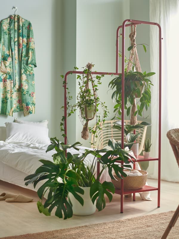 Une chambre à coucher avec de petites et de grandes plantes au bout du lit, suspendues et déposées sur un portant NIKKEBY rouge.