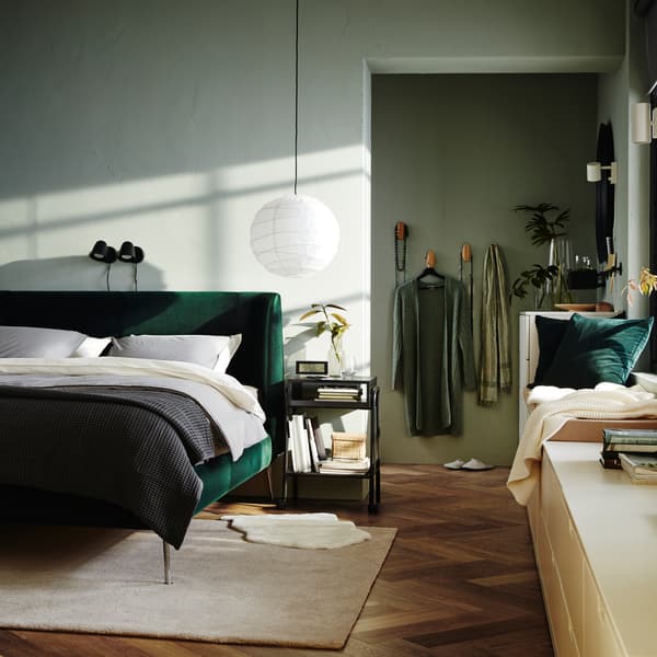 Une chambre à coucher avec un cadre de lit matelassé en velours vert, deux appliques noires, un tapis blanc cassé et une commode blanche.