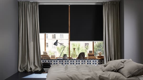 Une chambre à coucher avec un lit au centre de la pièce et des fenêtres entourées de rideaux INGERT et munies de stores à enrouleur occultants TRETUR à moitié baissés.