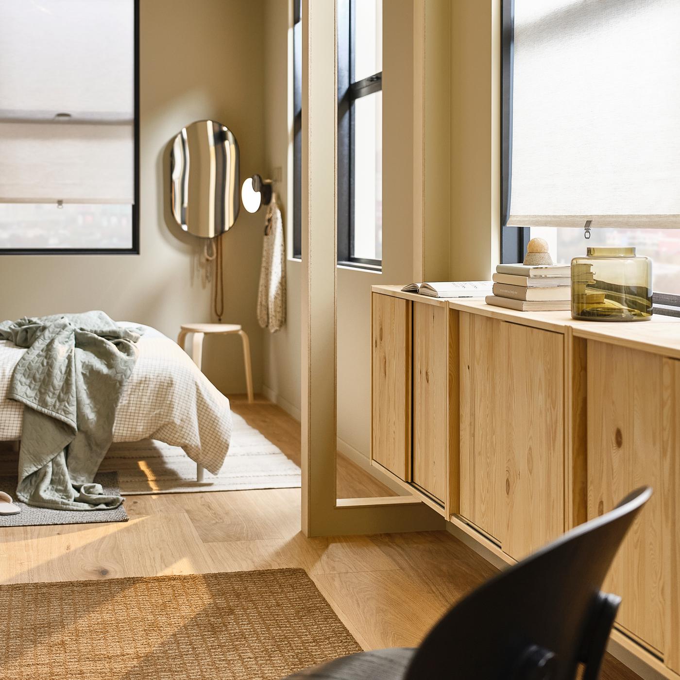 Une chambre à coucher avec un lit sur un tapis gris et un tapis beige, un miroir LINDBYN fixé au mur et des éléments IVAR en pin sous les fenêtres.