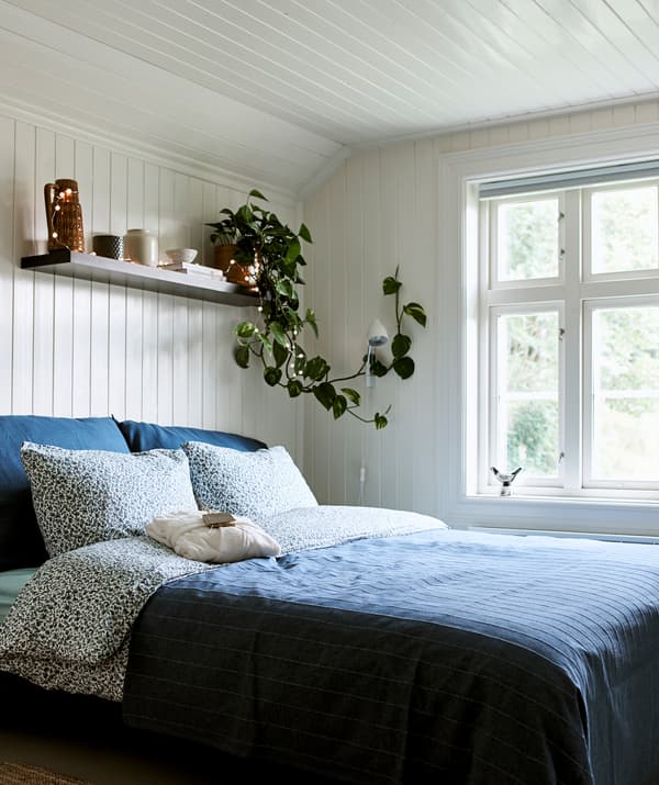 Une chambre à coucher dans une ferme avec des panneaux blanc en bois et un lit recouvert de linge de lit bleu avec une étagère murale au-dessus pour ranger et exposer.