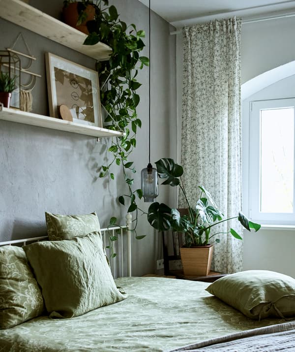 Une chambre à coucher décorée de textiles verts, de plantes et de deux étagères fixées sur un mur gris sur lesquelles est exposé un mélange d’arts et de petits objets.