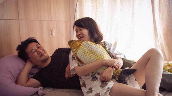 Une chambre à coucher illuminée et un jeune couple en train de rire et de se détendre sur le lit avec un coussin jaune en forme de poisson.