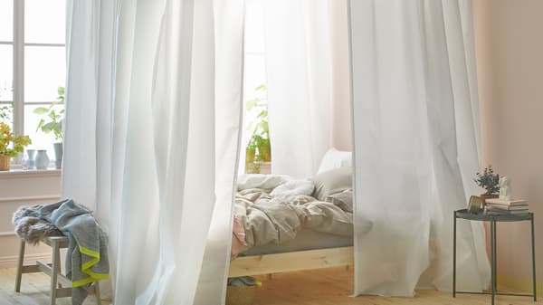 Une chambre à coucher lumineuse avec un lit près d’une fenêtre. Le lit est doté d’un ciel de lit fait à partir de rideaux blancs et de rails pour rideaux VIDGA.