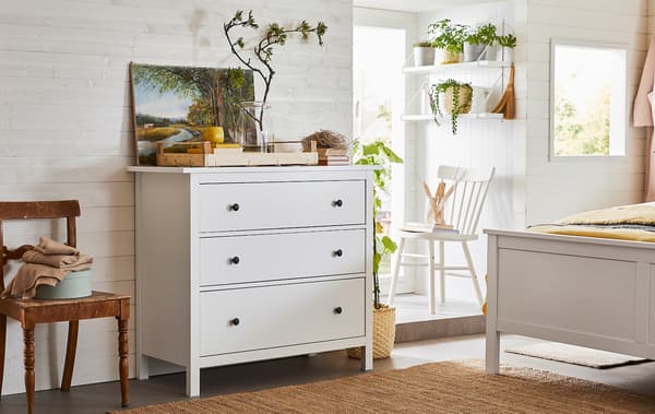 Une chambre bien aérée avec une prédominance d’articles en bois et de plantes vertes et une commode à trois tiroirs près de l’extrémité du lit.
