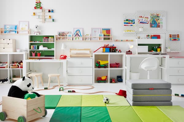 Une chambre d’enfant avec des jouets dispersés sur le sol, des bureaux sur le côté, des dessins sur des panneaux perforés et des rangements FLISAT sur les murs.