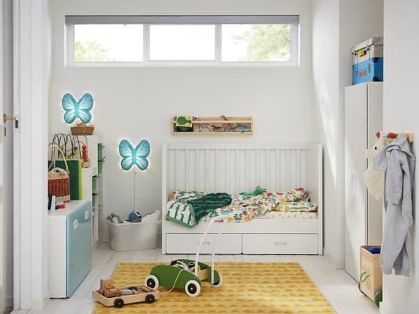 Une chambre d’enfant avec lit d’enfant, un tapis jaune, une armoire blanche, un chariot de marche vert et des appliques en forme de papillon.