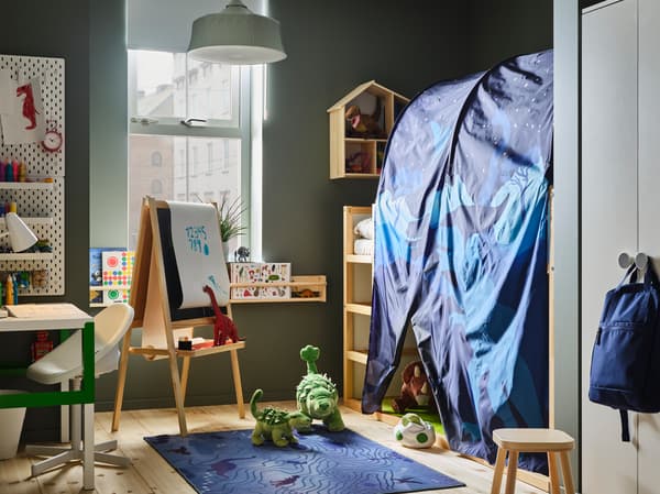Une chambre d’enfant avec un ciel de lit bleu, des dinosaures en peluche, un bureau blanc/vert, une suspension vert clair et un tapis bleu.