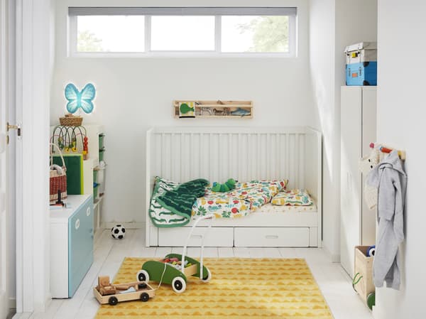 Une chambre d’enfant avec un lit enfant blanc, un tapis jaune, une armoire-penderie blanche, un porteur vert et une lampe murale en forme de papillon.