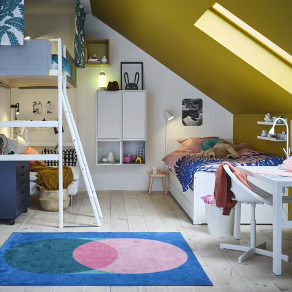 Une chambre d’enfant avec un lit mezzanine, un cadre de lit blanc, des armoires murales et rangements ouverts, un bureau blanc et un tapis rose/vert sur fond bleu.