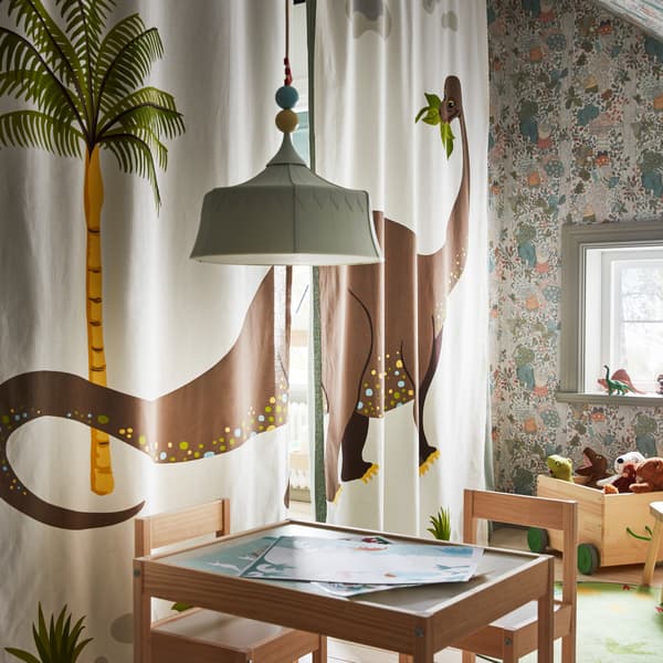 Une chambre d’enfant avec une table, des chaises et des jouets éparpillés.  Un des murs se compose de deux rideaux fermés ornés d’un motif de dinosaure. 