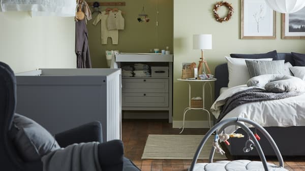 Une chambre pour parents et bébé avec un lit matelassé gris, un lit à barreaux gris, une commode/table à langer grise et des murs verts.