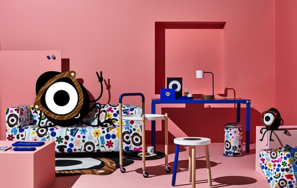 Une chambre rose avec un bureau bleu, une table d'appoint et un tabouret et un cyclope malicieux installé dans un canapé aux motifs multicolores.