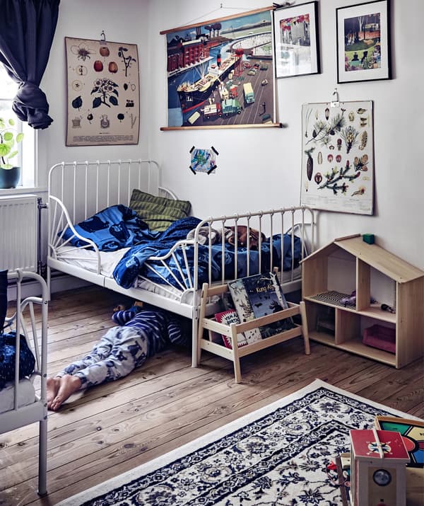 Une chouette chambre à coucher d’enfant avec des murs blancs, des rangements pour livres, une maison de poupée et de nombreux cadres au mur.