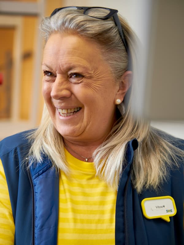 Une collaboratrice IKEA qui porte un chandail jaune et une veste bleue, souriant avec ses lunettes relevées sur la tête.