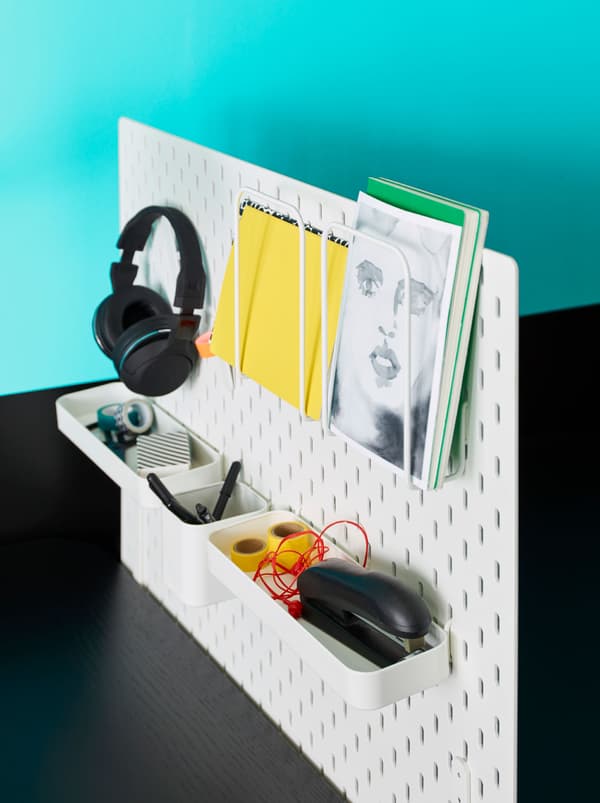 Une combinaison de panneau perforé blanc SKÅDIS avec tablette sur laquelle sont rangés du matériel de papeterie, un casque audio sur un crochet, et des documents dans des supports pour lettres.