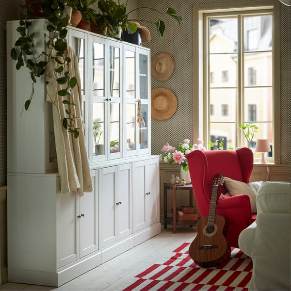 Une combinaison de rangement blanche avec des portes en verre dans une salle de séjour rouge et blanche. Des plantes vertes sont placées dessus.
