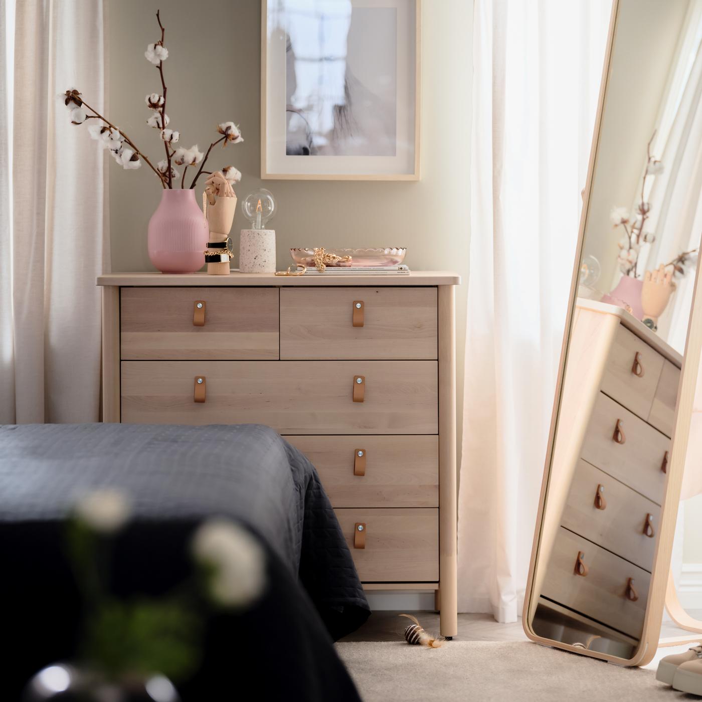 Une commode 5 tiroirs BJÖRKSNÄS en bouleau, avec des objets décoratifs sur le dessus, à côté d’un miroir sur pied IKORNNES dans une chambre.