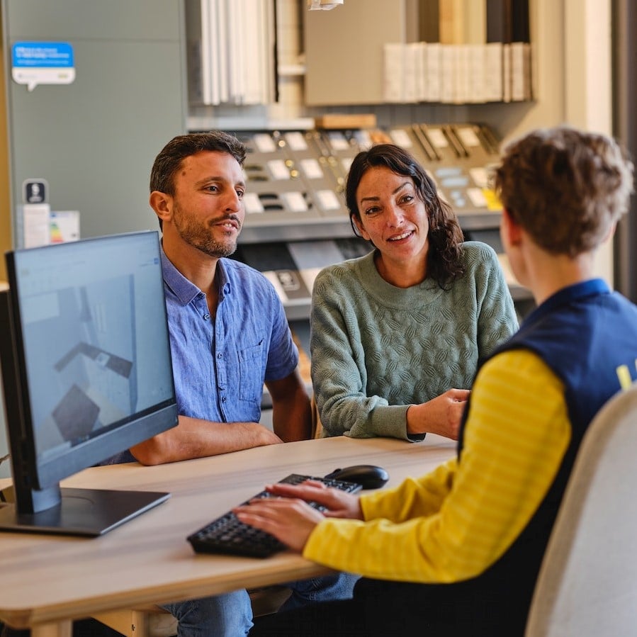 Une conseillère à la clientèle est assise à une table en face de deux clients, devant elle un écran sur lequel s’affiche un programme 3D avec une planification de cuisine.