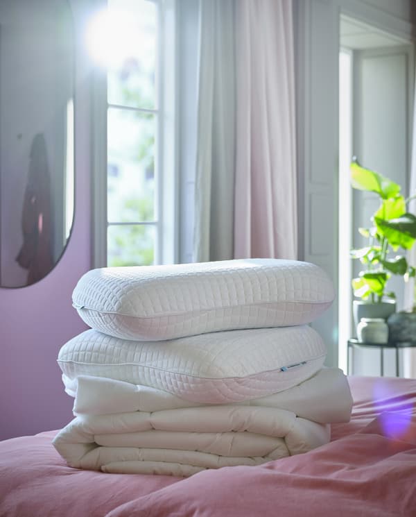 Une couette blanche et deux oreillers ergonomiques KLUBBSPORRE blancs empilés sur un lit avec linge de lit rose clair.