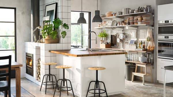 Une cuisine avec îlot blanc/bois brillant, une grille murale en acier inoxydable avec des étagères, et des tabourets en pin/noir.