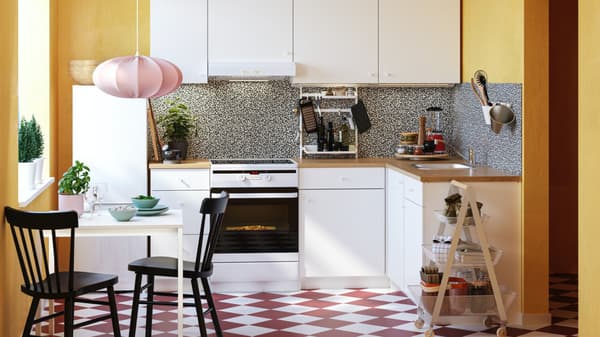 Une cuisine blanche dans une pièce jaune avec un sol à carreaux blancs et rouges et des chaises noires à une petite table avec une lampe suspendue rose