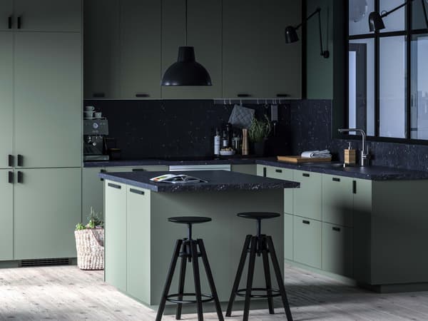 Une cuisine BODARP gris-vert avec un îlot, des comptoirs effet marbré noir, des tabourets de bar noirs et une grande plante près de la fenêtre.