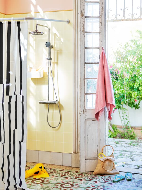 Une douche ouverte avec un rideau de douche noir et blanc, à côté d’une porte ouverte en bois blanc patiné.