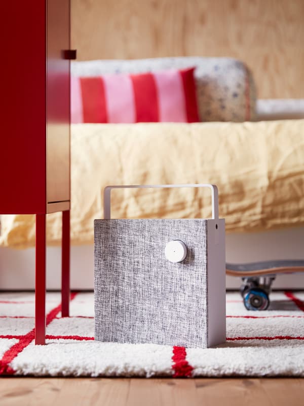 Une enceinte bluetooth ENEBY blanche sur un tapis rouge et blanc dans une chambre à coucher moderne, lumineuse, aux murs revêtus de panneaux de bois.