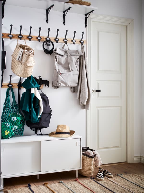 Une entrée avec des crochets auxquels sont suspendus des vestes et des sacs, une étagère large placée en hauteur et un banc MACKAPÄR blanc avec des rangements.
