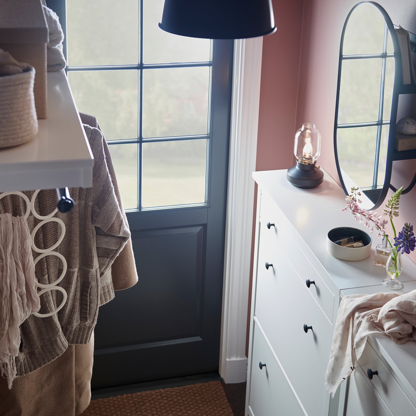 Une entrée avec deux range-chaussures blancs sur lesquels sont posés divers articles, une étagère-patère blanche et un miroir avec rangement noir.