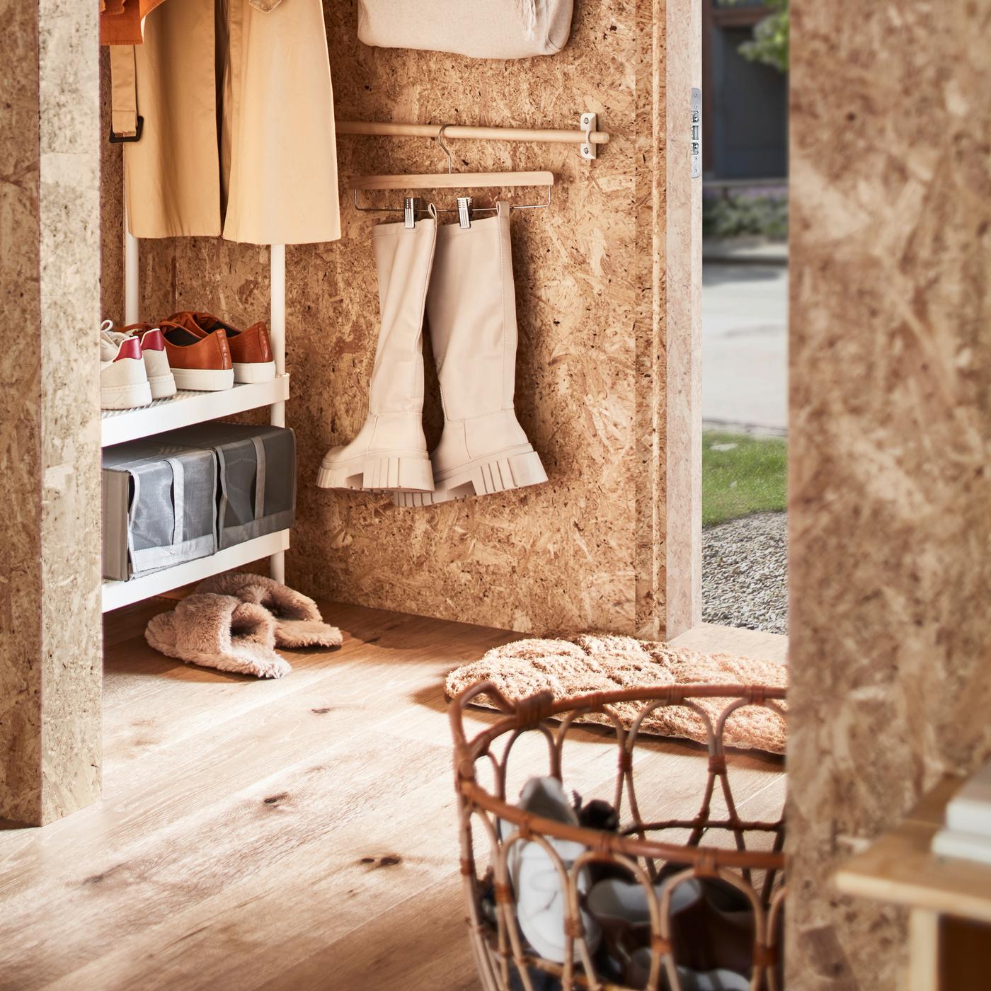 Une entrée avec un rangement ouvert pour vêtements et chaussures, un panier en rotin contenant des chaussures et une barre supportant des bottes sur un porte-jupe.