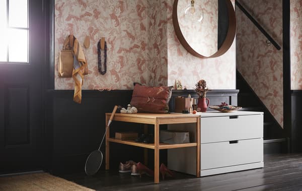 Une entrée avec une commode deux tiroirs NORDLI gris clair placée sous un miroir rond et à côté d’un banc en bois avec des étagères ouvertes.