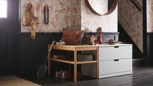 Une entrée avec une commode deux tiroirs NORDLI placée sous un miroir rond et à côté d’un banc en bois avec des étagères ouvertes.