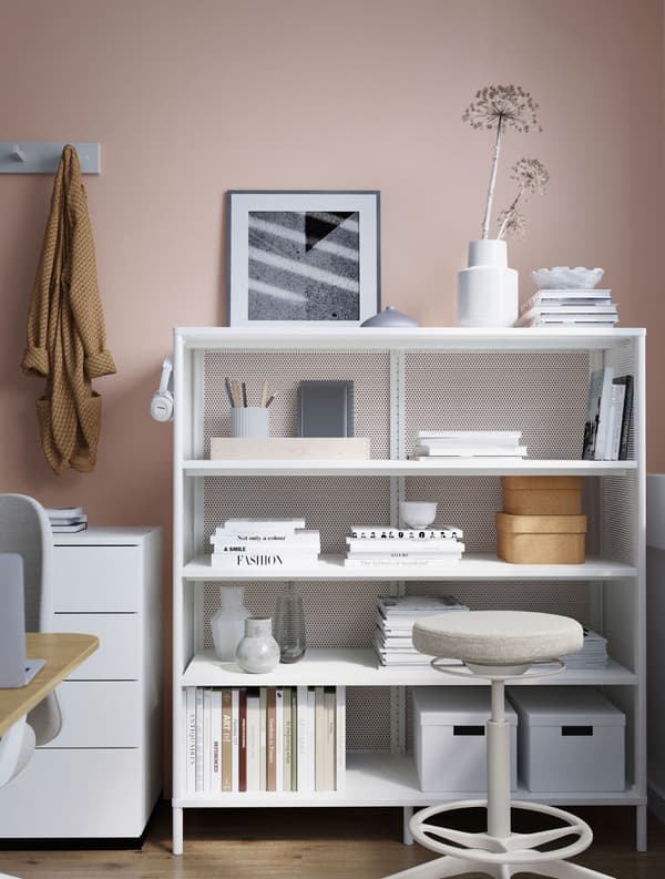 Une étagère BEKANT blanche contenant des livres, des vases, du matériel de papeterie, un casque et des boîtes de rangement. Une chaise couleur crème est installée devant l’étagère.