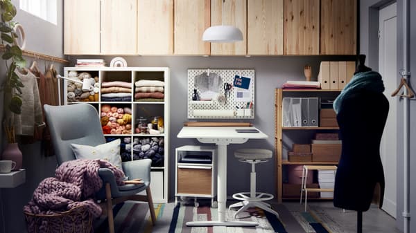 Une étagère blanche avec des pelotes, un fauteuil vert clair, un bureau avec structure assis/debout blanc, des rangements en pin et une suspension.
