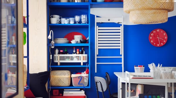 Une étagère PLATSA bleue est fixée sur un mur bleu, près d’une chaise pliante FRÖSVI blanche suspendue à des crochets.