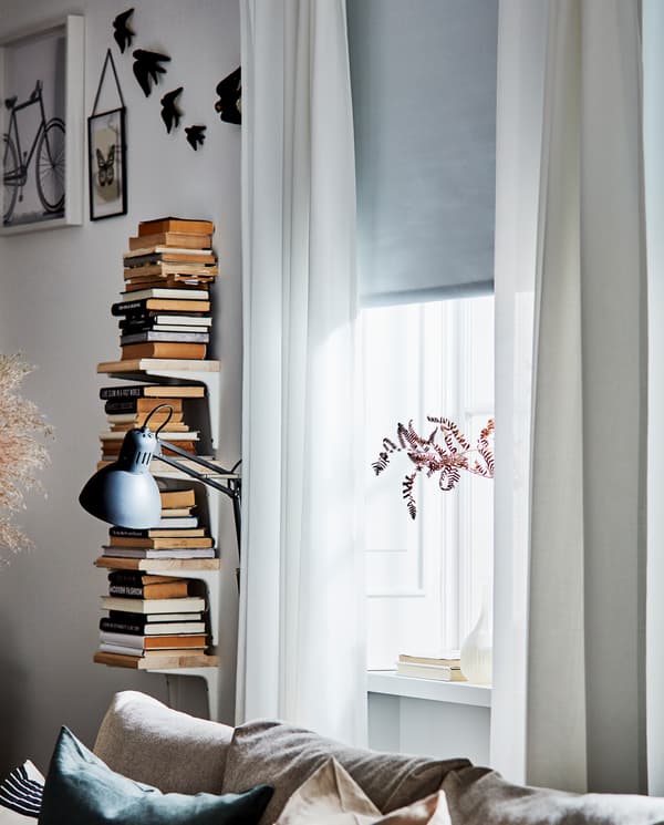 Une étagère verticale blanche en bois avec des livres, des rideaux blancs et un store à enrouleur opaque blanc qui couvre la moitié de la fenêtre.