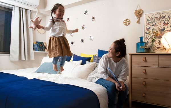 Une femme assise au bord d’un grand lit sur lequel saute une petite fille dans une chambre blanche avec une étagère à livres.