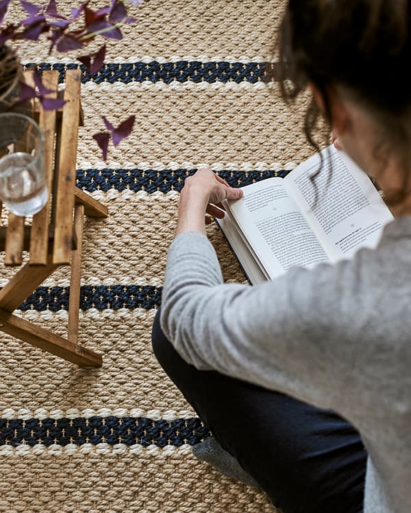 Une femme assise lit sur un tapis rayé tissé dans des fibres naturelles avec une table pliable, des plantes et un verre d’eau.