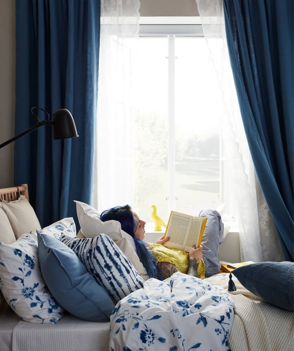 Une femme assise sur un lit et lisant un livre; son pied sur l’appui de fenêtre, elle se repose sur une montagne de coussins.