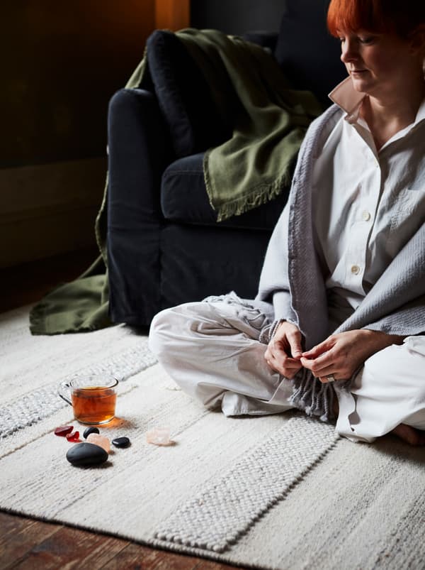 Une femme assise sur un tapis de couleur claire dans un séjour; des cristaux, des pierres et une tasse en verre sont posés sur le sol devant elle.
