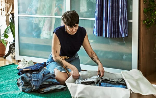 Une femme aux bras tatoués est assise sur un tapis vert dans une chambre à coucher; elle trie une pile de jeans dans un sac de rangement en tissu couleur crème.