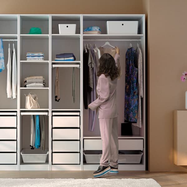 Une femme aux cheveux foncés suspend une robe sur la tringle à habits d’une combinaison d’armoire-penderie ouverte PAX blanche, dans une pièce aux murs bruns.
